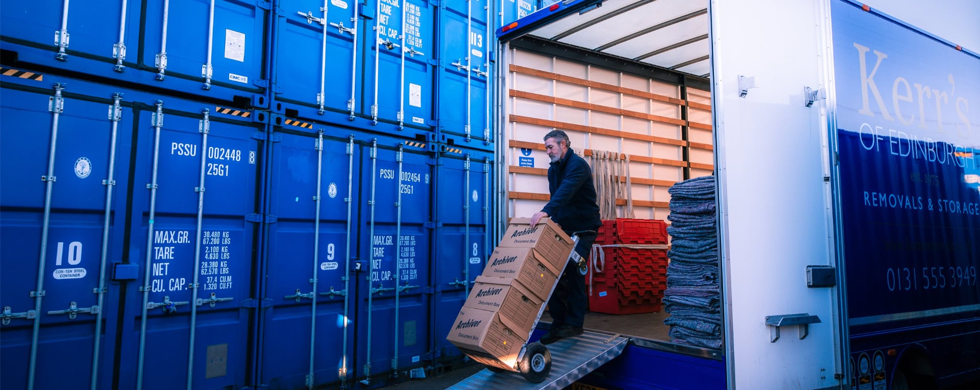 Staff member unloading archive boxes into blue container storage units beside a Kerr’s Removals van in Edinburgh